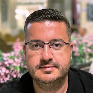 A man with short dark hair and glasses, wearing a black shirt, sits indoors with pink flowers and blurred lights in the background.