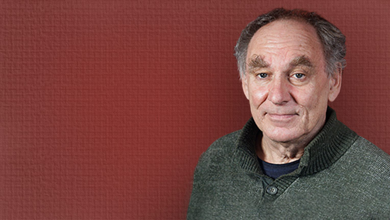 An older man with grey hair and a neutral expression wears a green jumper and stands in front of a plain reddish-brown background.