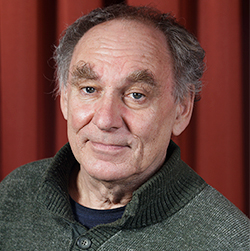 An older man with grey hair and thick eyebrows wears a green knitted jumper and smiles slightly in front of a red curtain background.