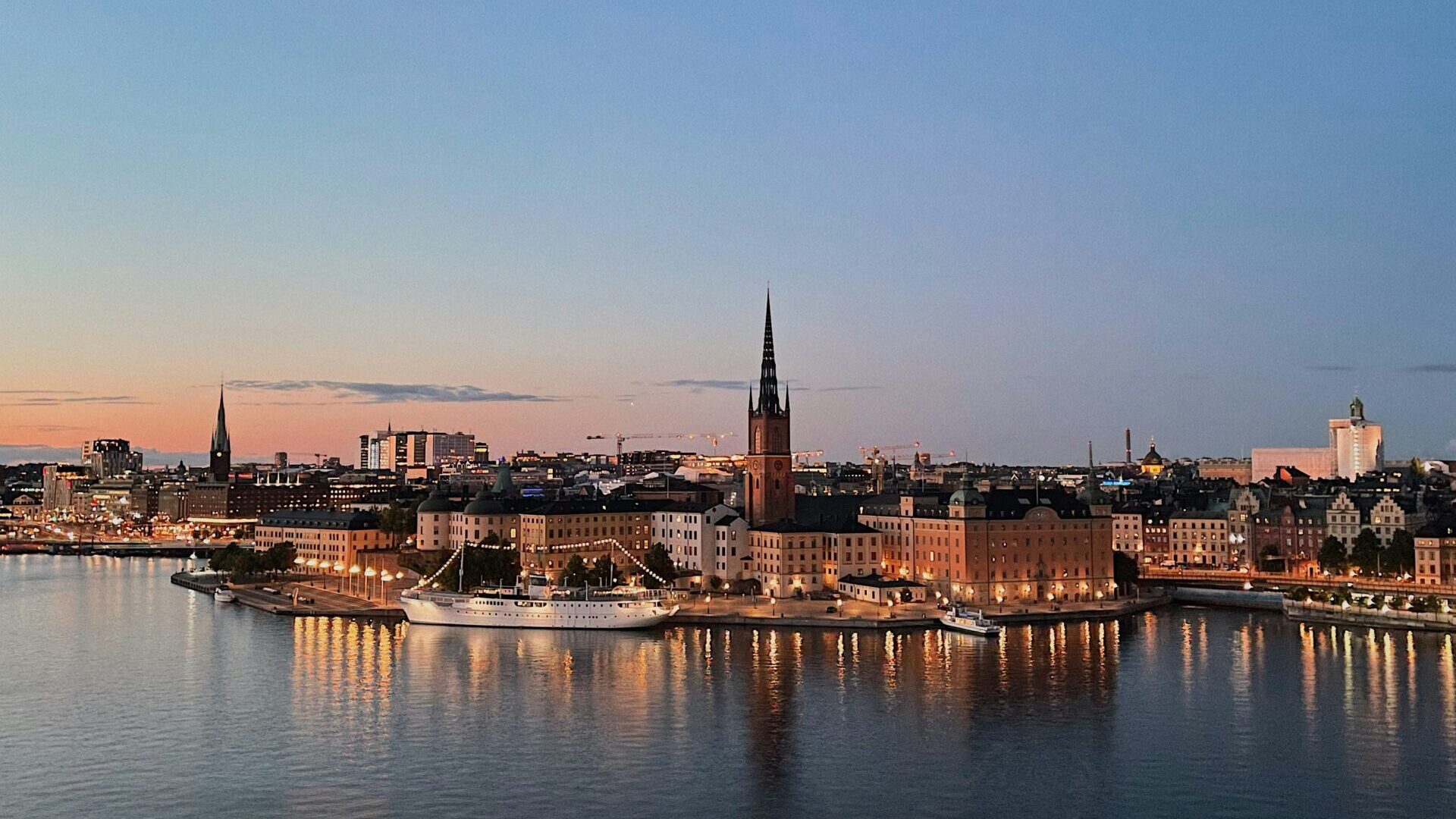 Cityscape at dusk showing Stockholm’s waterfront buildings with warm lights reflecting on the calm water. A prominent church spire rises in the centre against a clear blue sky. Trees frame the view in the foreground.
