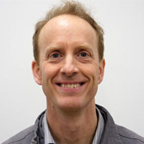 A middle-aged man with short light brown hair smiles at the camera. He is wearing a light grey collared shirt under a grey jacket, and is posed against a plain white background.