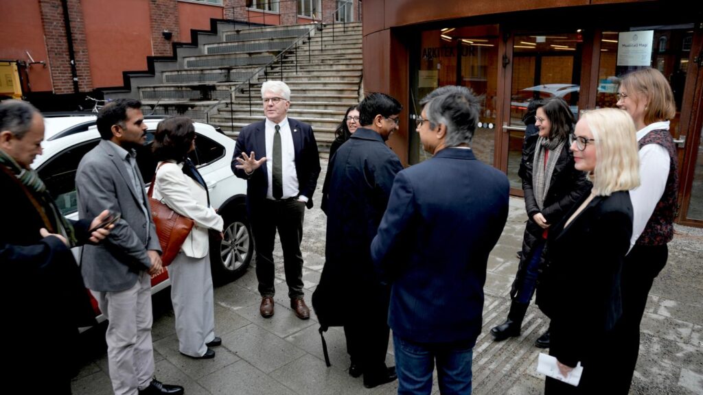 A group of people stand in a circle outdoors on a paved area, talking and listening to a man in a suit who gestures with his hand. Buildings and stairs are visible in the background.
