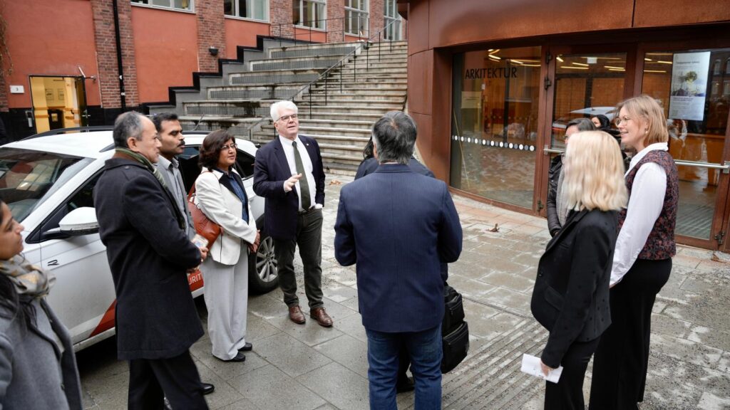 A group of people, including men in suits and women in business attire, stand talking outside a modern brick and glass building near a white car on a city pavement.