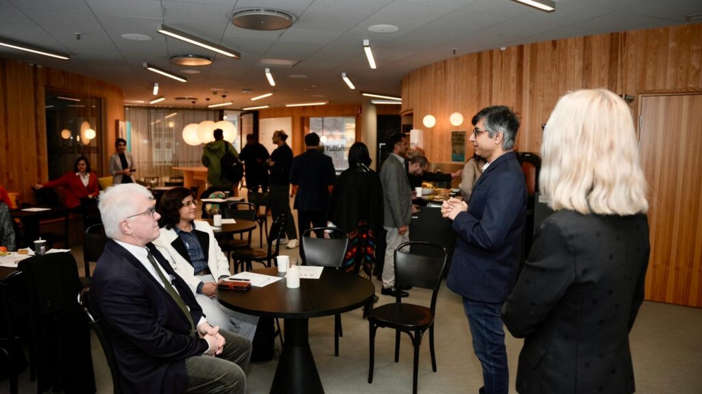 A group of people gather in a modern, wood-panelled room. Some are seated at round tables, while others stand and talk. The room is warmly lit with hanging lamps. A man stands and speaks to the group.