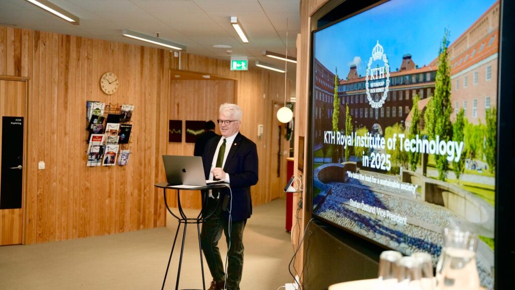A man in a suit stands at a small table with a laptop, giving a presentation in a modern, wood-panelled room. A large screen displays KTH Royal Institute of Technology in 2025 with campus buildings in the background.