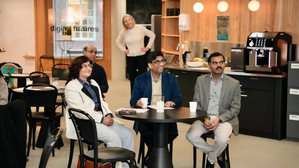 Four people sit at a round table, smiling and looking forward in a modern café setting, whilst a woman in the background stands and smiles near a kitchen area with a coffee machine.