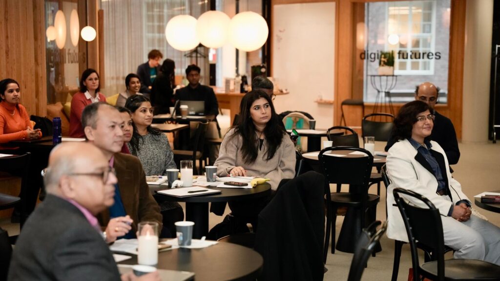 A diverse group of people sit attentively at tables in a modern, well-lit room with large round lights and glass walls, listening to a presentation at an event or workshop.