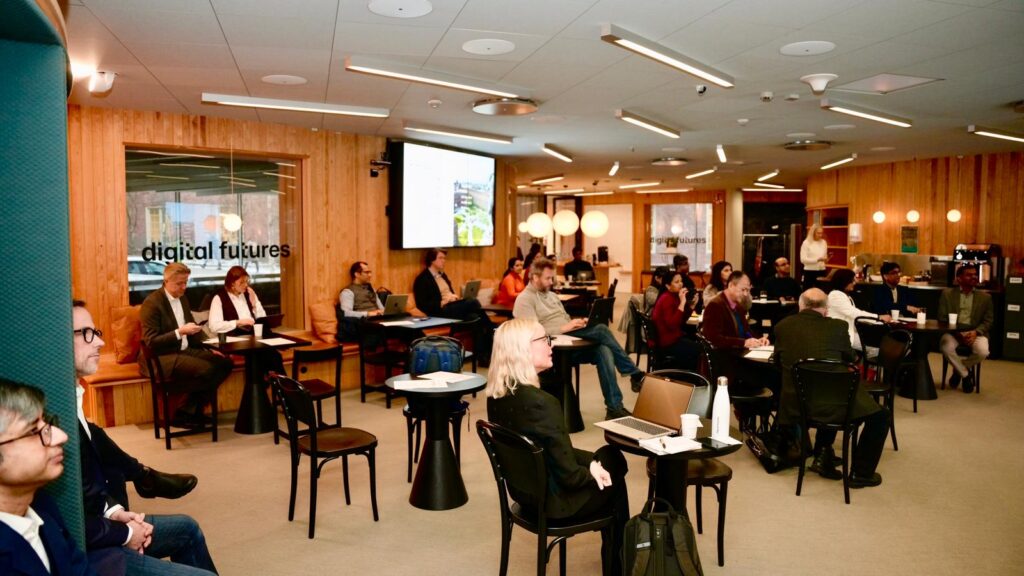 A group of people sit at tables in a modern, well-lit conference room with wood panelling. A screen displays a presentation, and a glass wall with the words digital futures is visible in the background.