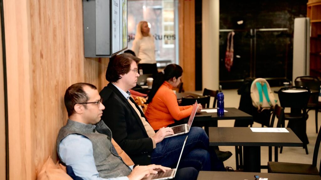 Several people sit at tables with laptops in a modern, wood-panelled room, focusing on their screens. A woman stands at the front, possibly presenting or teaching. Rucksacks and papers are visible on the tables.