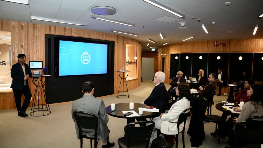 A man stands and presents at the front of a modern conference room with a large screen displaying a blue slide. Several people sit at tables, listening attentively. The room has wood-panelled walls and a professional atmosphere.
