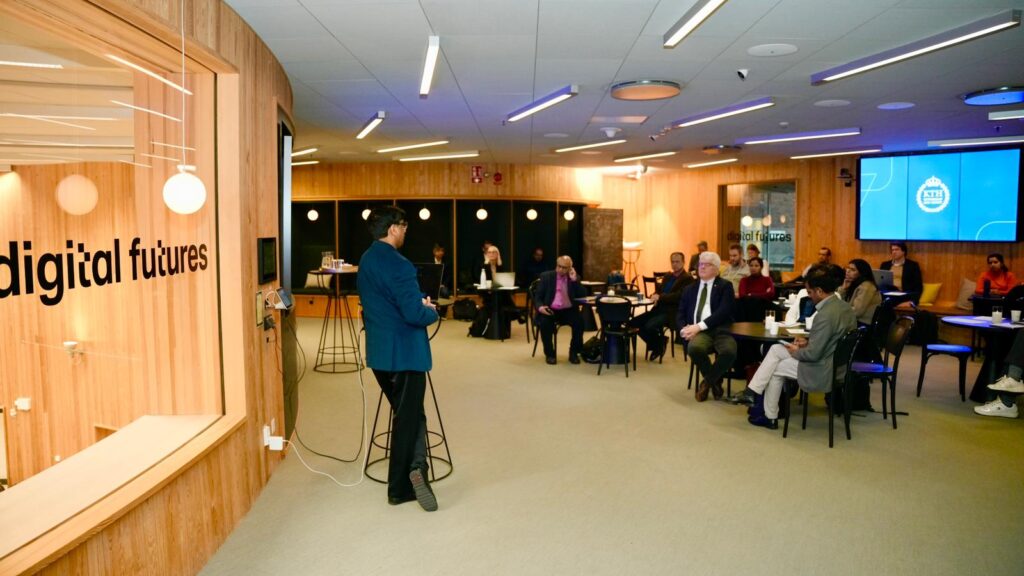 A person stands and speaks to a group of people seated at tables in a modern, well-lit room with wood-panelled walls. A glass window reads digital futures, and screens display blue graphics.
