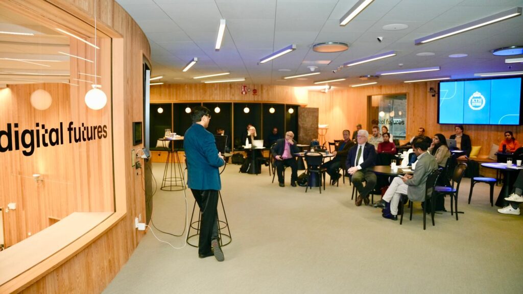 A person speaks to an audience seated at tables in a modern, wood-panelled room with a sign reading digital futures on the wall and presentation slides displayed on screens.
