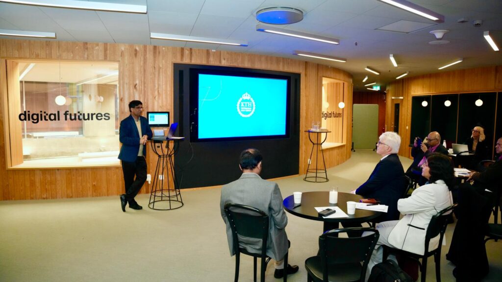 A presenter stands by a screen displaying a blue slide in a modern conference room labelled digital futures, speaking to a seated audience of professionals.