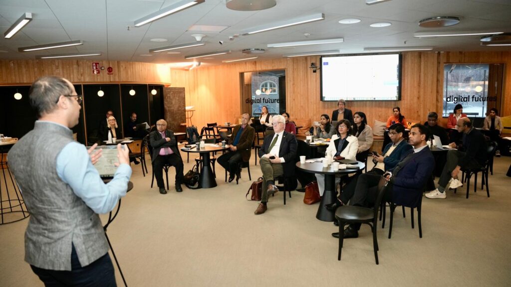A man speaks to a group of people seated at small tables in a modern conference room with wood-panelled walls and large windows, whilst a presentation screen is visible in the background.