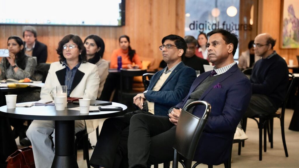 A group of professionals sit attentively at tables during a seminar or conference in a modern indoor setting, with digital futures text visible on the wall in the background.