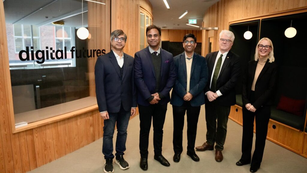 Five professionally dressed people stand in a corridor with wood-panelled walls and a glass window displaying the words “digital futures.” They are smiling and facing the camera.