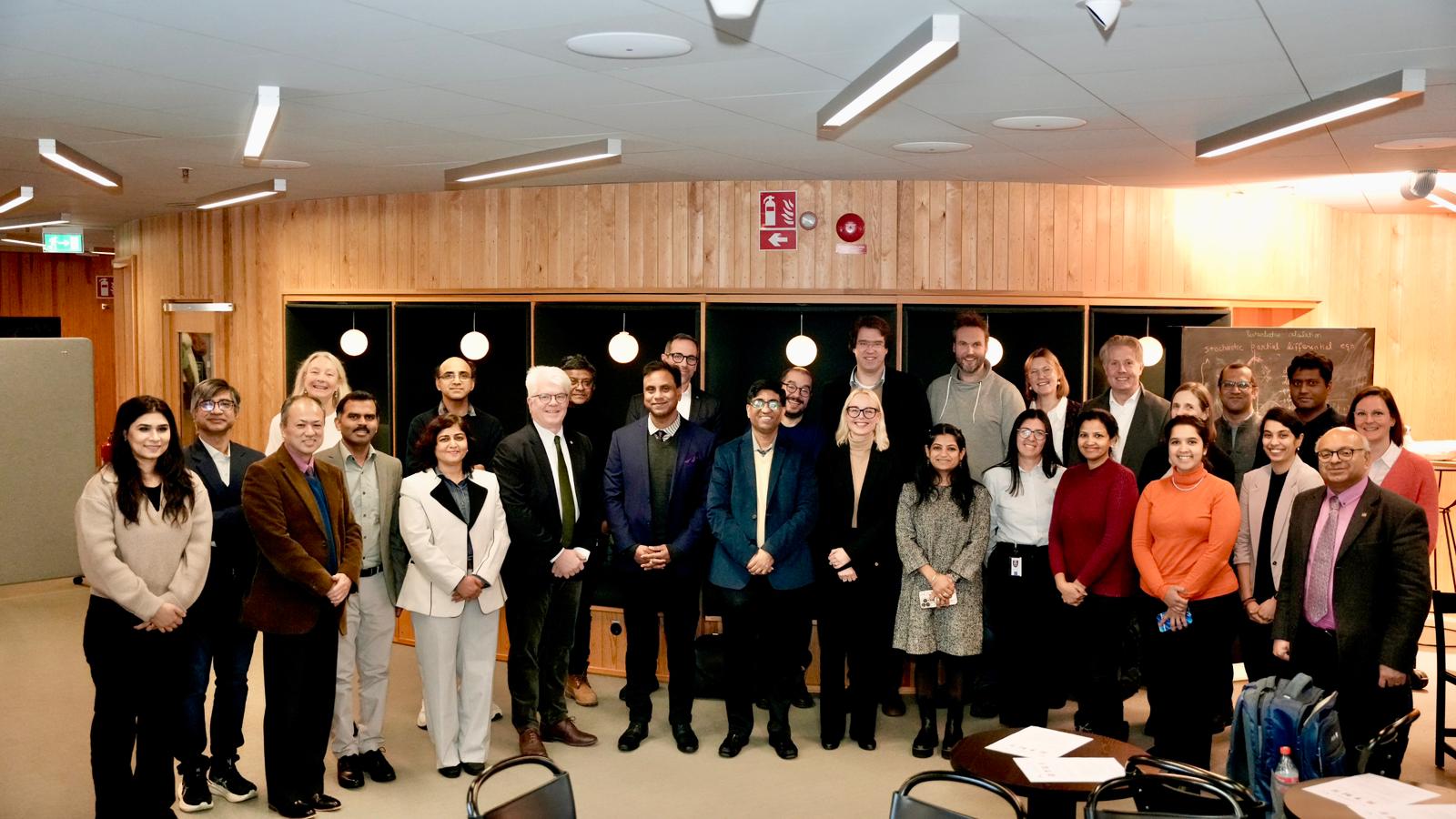 A large group of professionally dressed people stand together and smile for a group photo in a modern, well-lit indoor setting with wooden walls and round lights in the background.