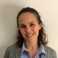 A woman with wavy brown hair smiles at the camera. She is wearing a light blue blouse and a grey blazer, standing against a plain off-white background.