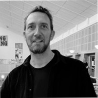 A man with short hair and a beard smiles while standing indoors in a spacious, well-lit room with high ceilings and wall decorations. The image is in black and white.