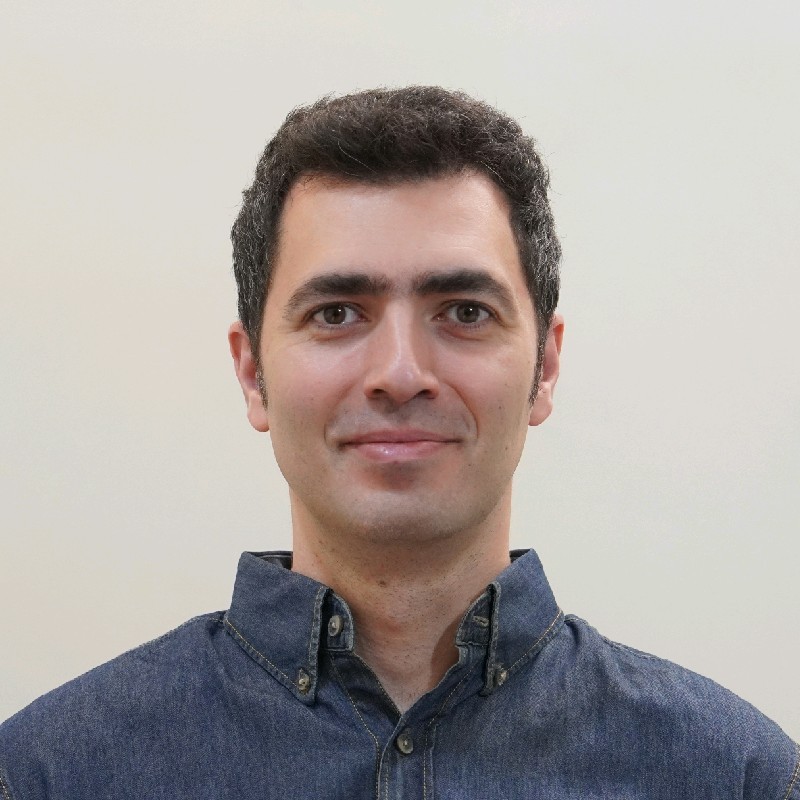 A man with short dark hair and fair skin, wearing a dark denim button-up shirt, is smiling softly while facing the camera against a plain light-coloured background.