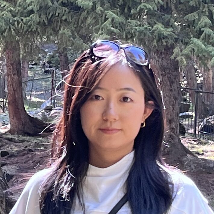 A woman with long dark hair wearing a white top and sunglasses on her head stands outdoors in a sunlit forested area with tall trees in the background.