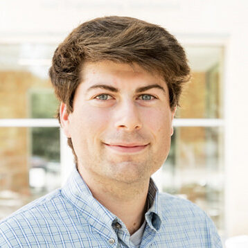 A young man with light skin and brown hair, wearing a blue checked button-up shirt, stands indoors with soft natural light and large windows in the background. An orange FLIP icon is at the bottom right corner.