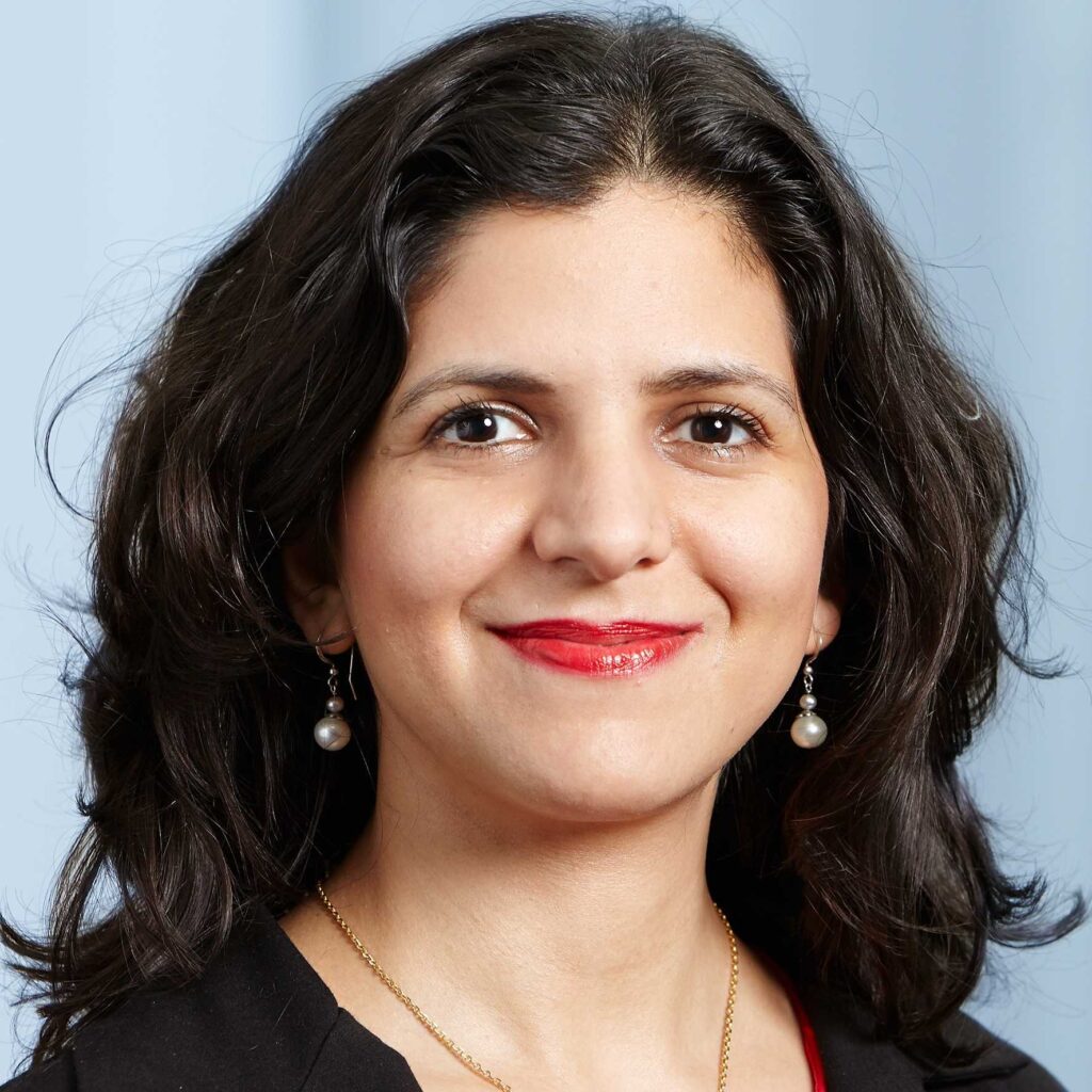 A woman with wavy dark hair, wearing red lipstick, pearl drop earrings, a gold necklace, and a black top, smiles at the camera against a light blue background.
