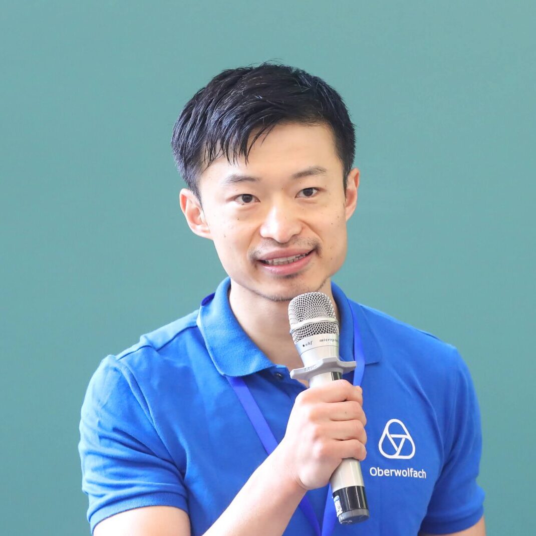A man in a blue polo shirt stands in front of a green chalkboard, holding a microphone and a remote, appearing to give a presentation. A conference badge hangs from his neck.