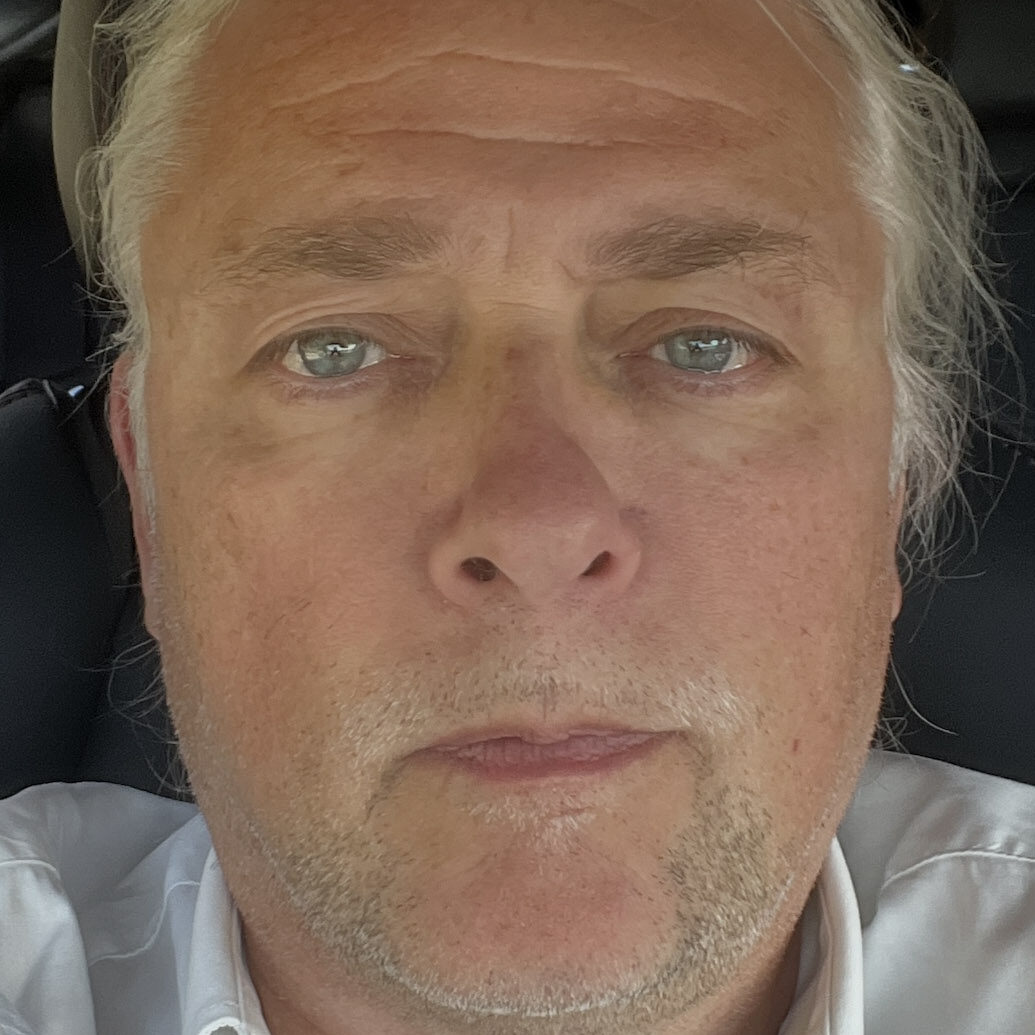 A middle-aged person with light skin, grey hair, and blue eyes looks directly at the camera. They are wearing a white collared shirt and are seated indoors, possibly in a car.