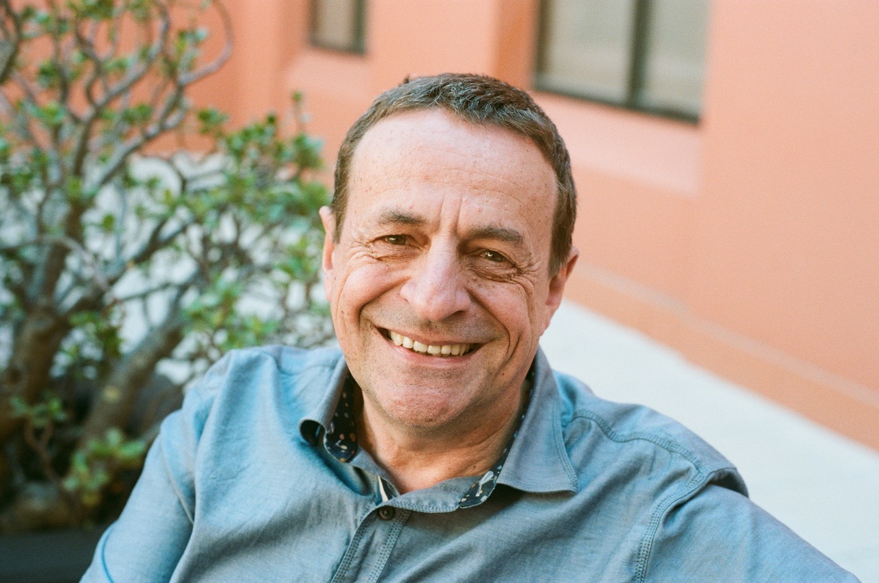 A middle-aged man with short brown hair and a blue button-up shirt smiles warmly at the camera whilst sitting outdoors near a plant and a peach-coloured building.