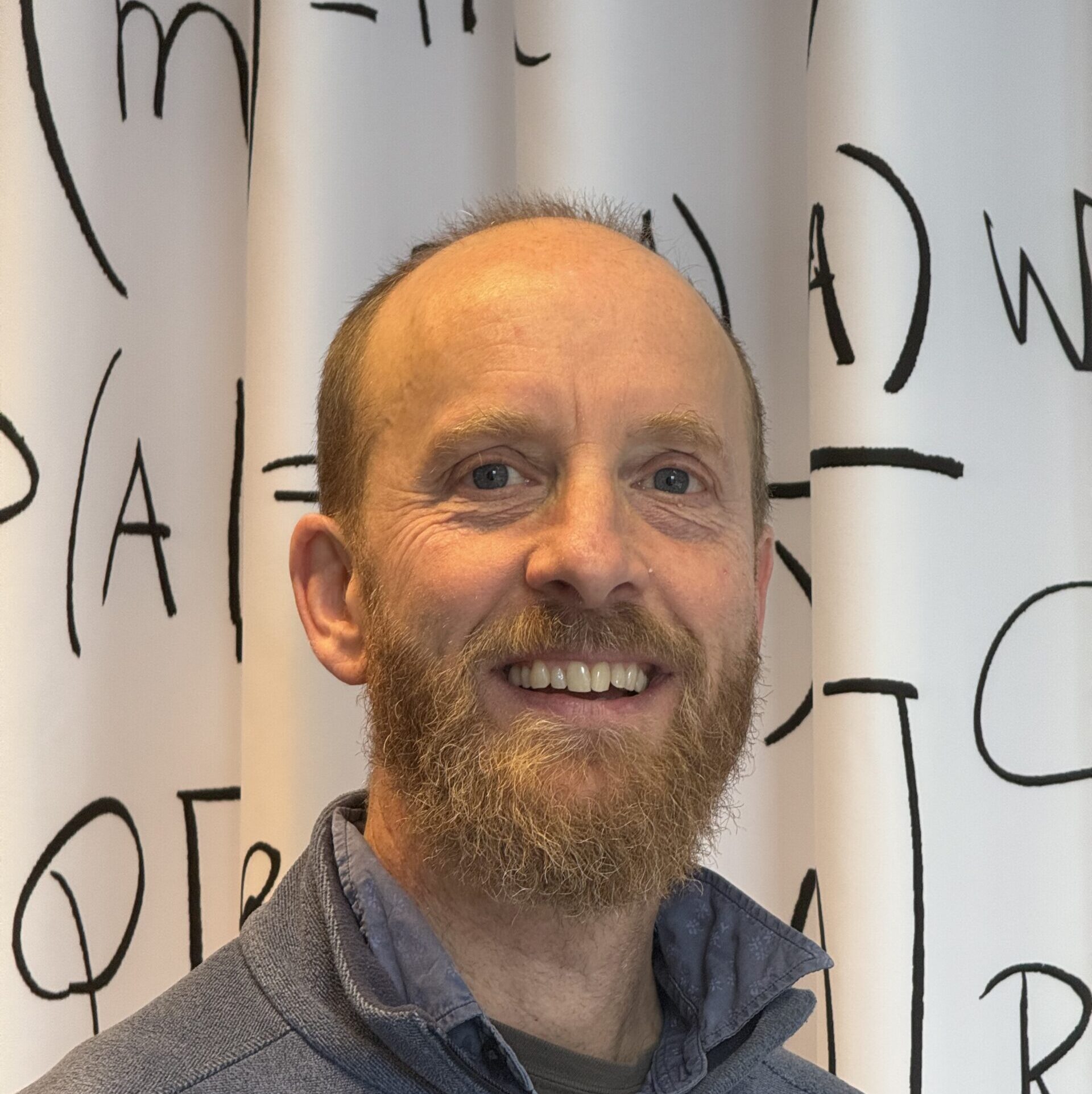 A smiling man with a beard and short hair, wearing a blue zip-up jacket, stands in front of a white curtain with black abstract letters and symbols.