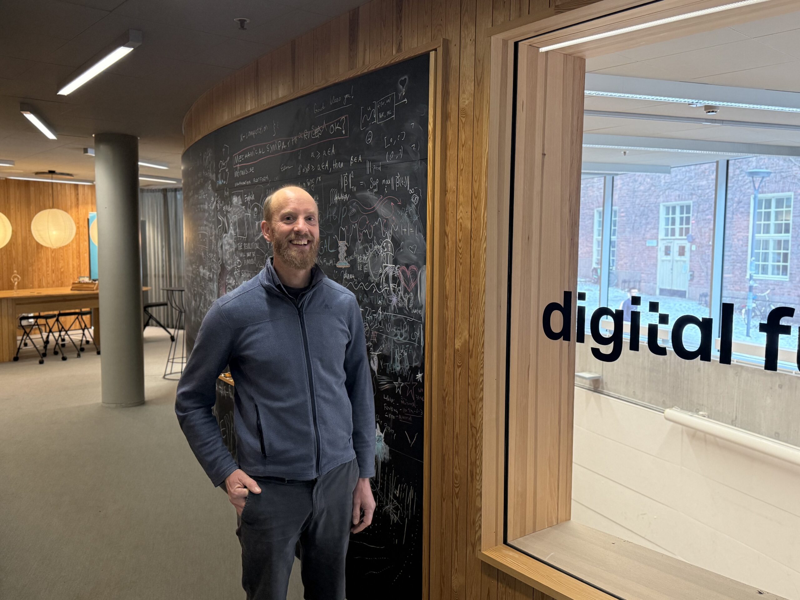 A smiling man stands in a modern office near a blackboard covered in chalk drawings and equations. Wooden walls and large windows create a bright, open environment. The partial words “digital f” appear on the window.