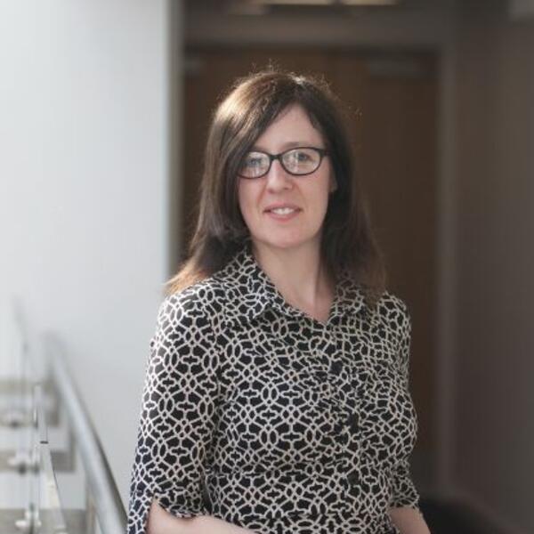 A woman with straight brown hair and glasses stands indoors, wearing a patterned black and white blouse. She is smiling softly and looking at the camera, with a blurred corridor background behind her.