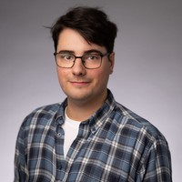 A person with short dark hair and glasses wearing a blue checked shirt over a white T-shirt, standing in front of a plain grey background and smiling slightly.