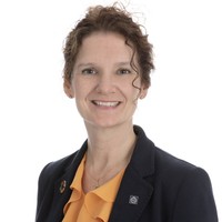 A woman with curly brown hair, wearing a navy blazer over a yellow blouse, smiles at the camera against a plain white background.