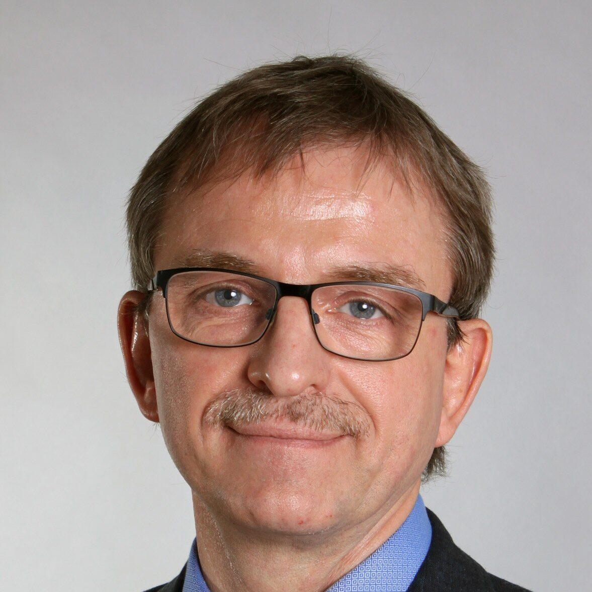 A middle-aged man with short light brown hair, glasses, and a moustache, wearing a blue dress shirt, patterned blue tie, and a dark suit jacket, poses against a plain light grey background.