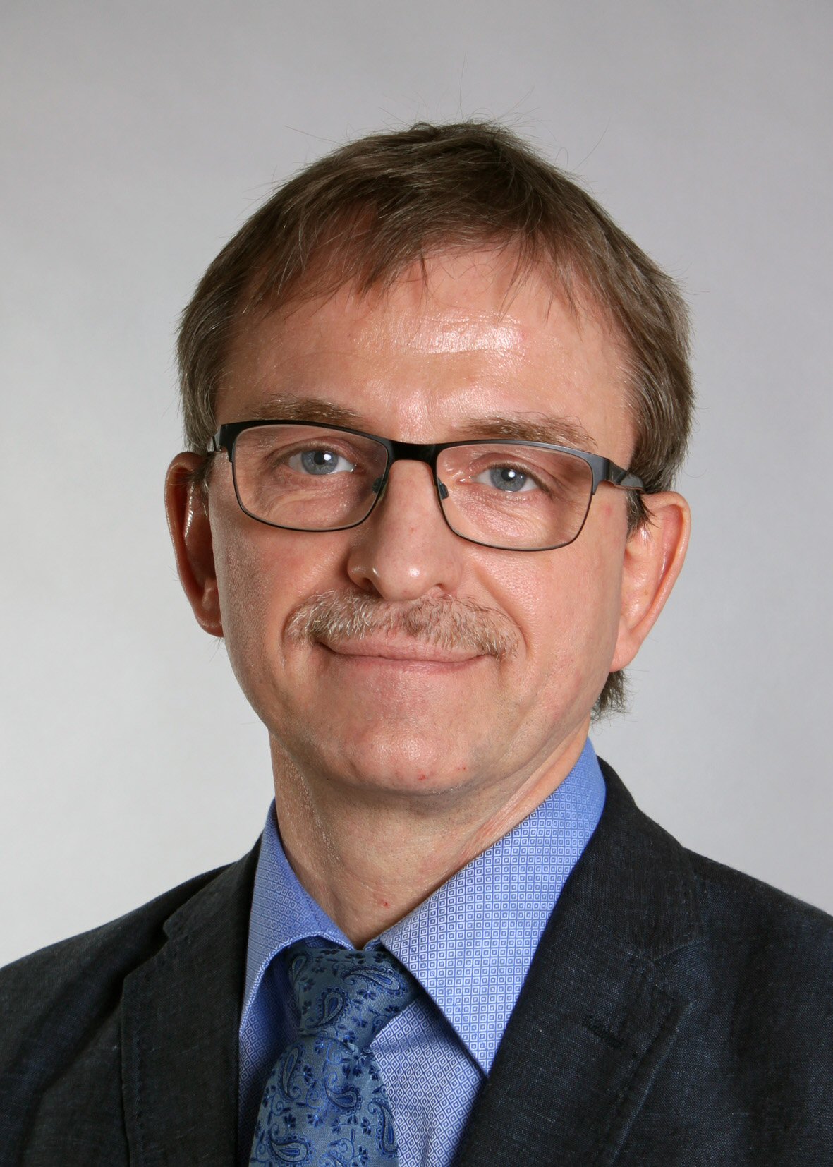 A middle-aged man with short light brown hair, glasses, and a moustache, wearing a blue dress shirt, patterned blue tie, and a dark suit jacket, poses against a plain light grey background.