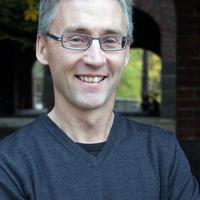 A middle-aged man with short grey hair and glasses is smiling, wearing a dark long-sleeved shirt, standing outside with brick arches and trees in the blurred background.