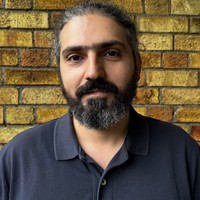 A man with dark hair tied back and a full beard is wearing a dark polo shirt, standing in front of a textured yellow and brown brick wall.