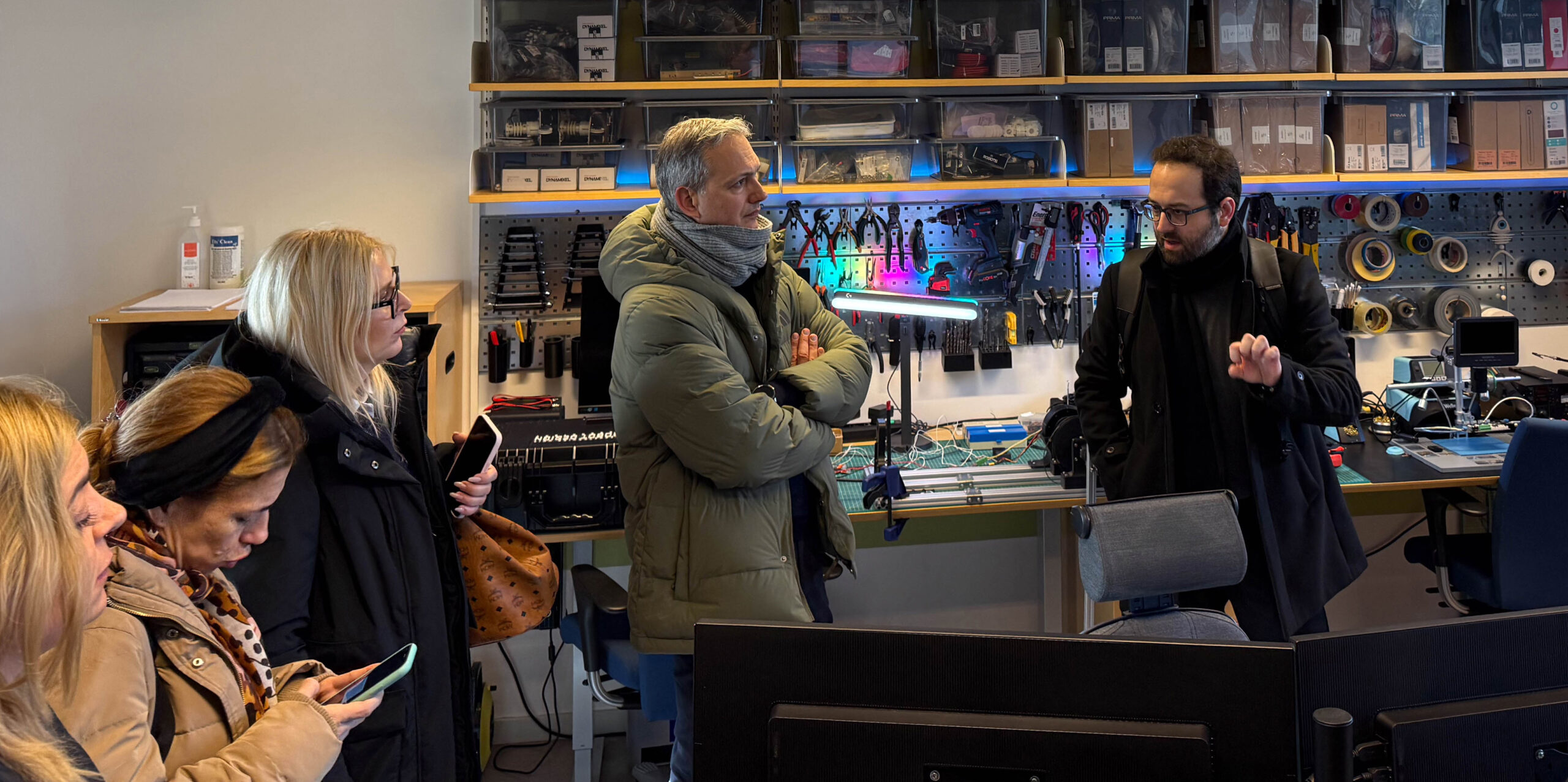 A group of people stands and talks in a workshop filled with electronics, tools, and equipment on shelves and benches. Some listen attentively whilst others check their mobiles.