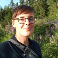 A person with short brown hair and glasses smiles outdoors in a sunlit area with green trees and purple wildflowers in the background.