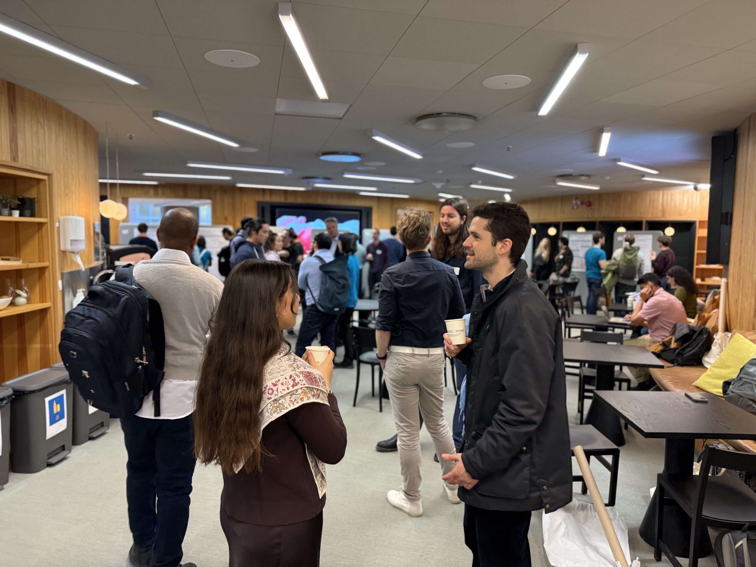 A group of people are standing and talking in a modern, well-lit room with wooden walls. Some hold cups and wear rucksacks. Several tables and chairs are scattered around, and a presentation screen is visible in the background.