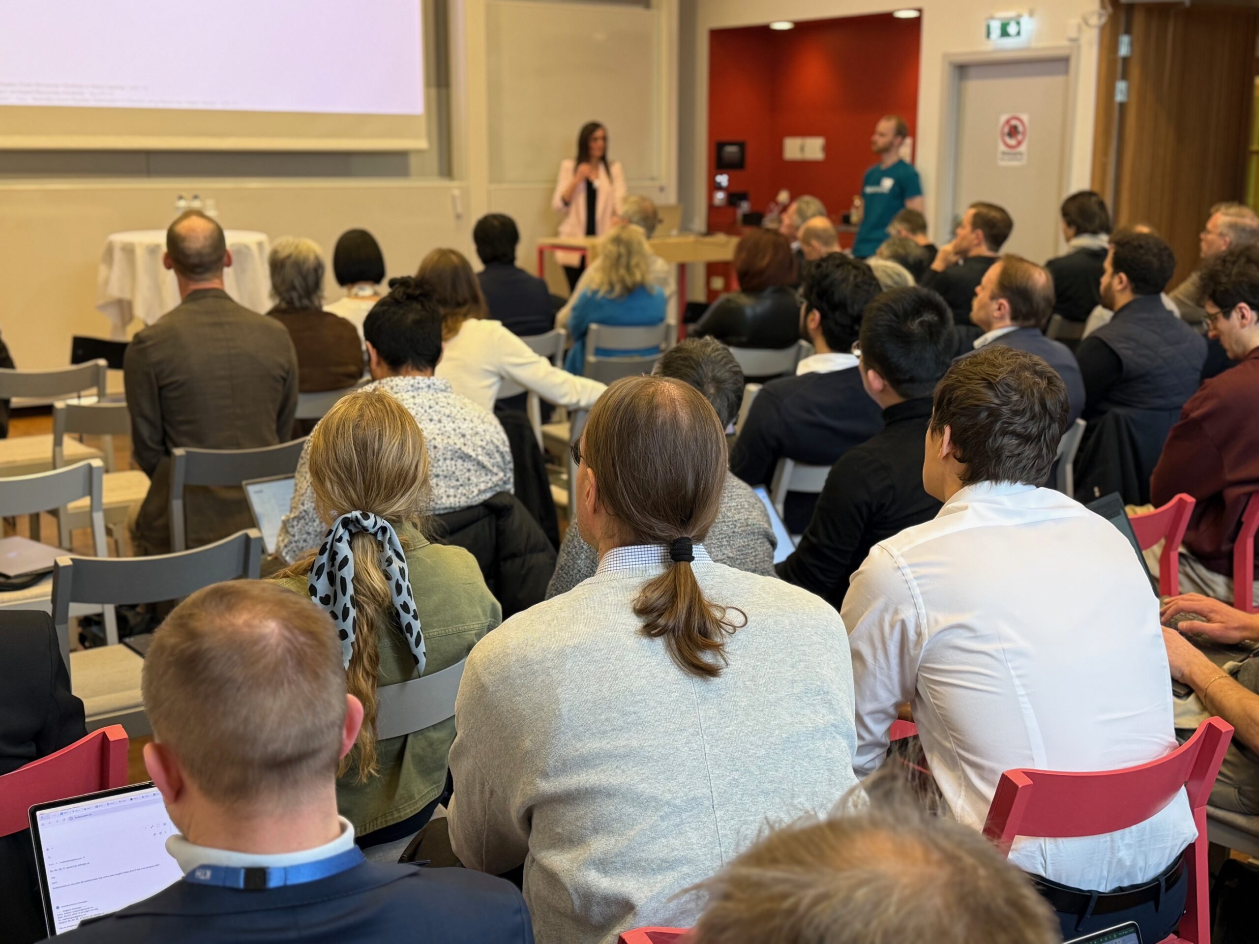 A group of people sit facing a woman speaking at a lectern in a classroom or conference room, with a man standing nearby and a projection screen behind them.