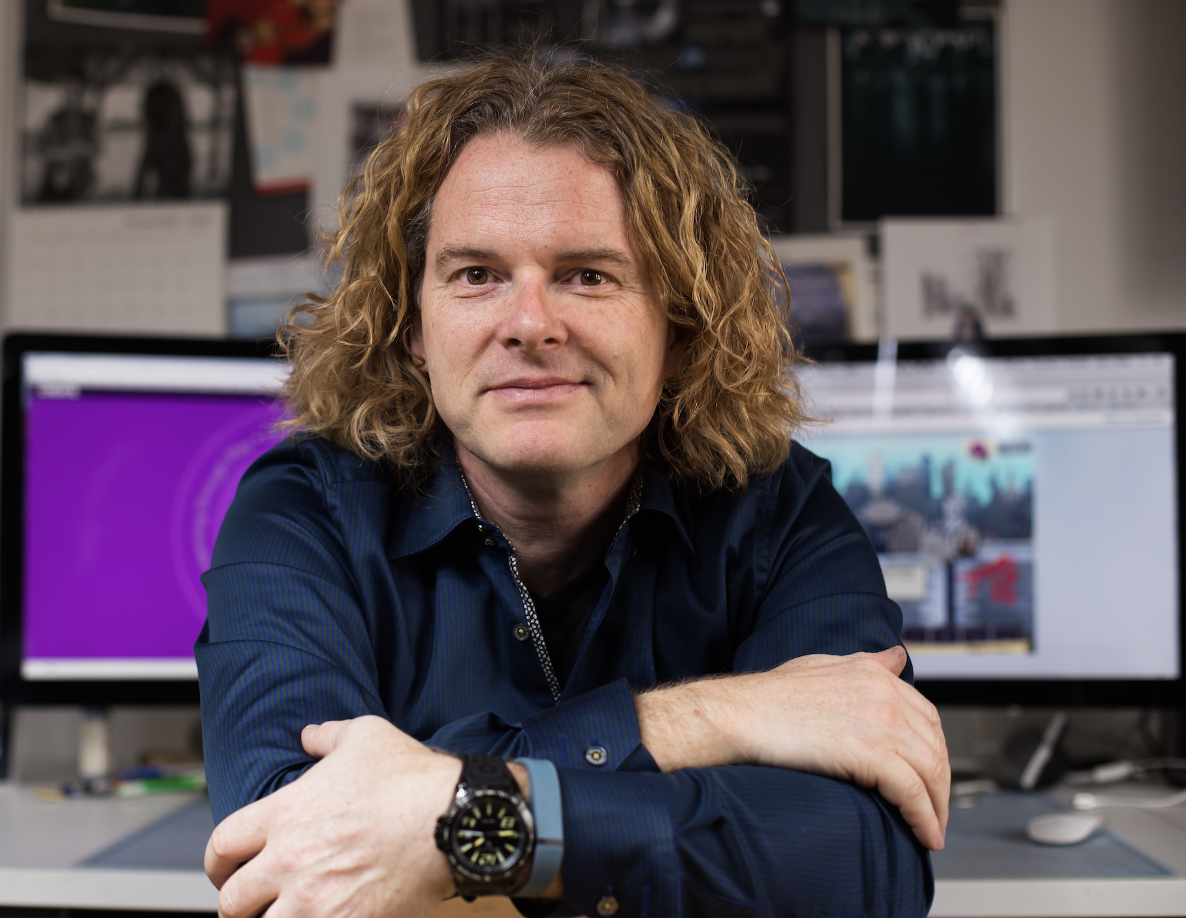 A person with curly, shoulder-length hair and a dark blue shirt sits with arms folded in front of two computer monitors in an office with papers and photos on the wall behind them.