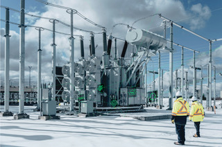 Two workers in safety gear walk towards large electrical equipment at an outdoor power substation, surrounded by metal structures, under a cloudy sky. Snow covers the ground and equipment.