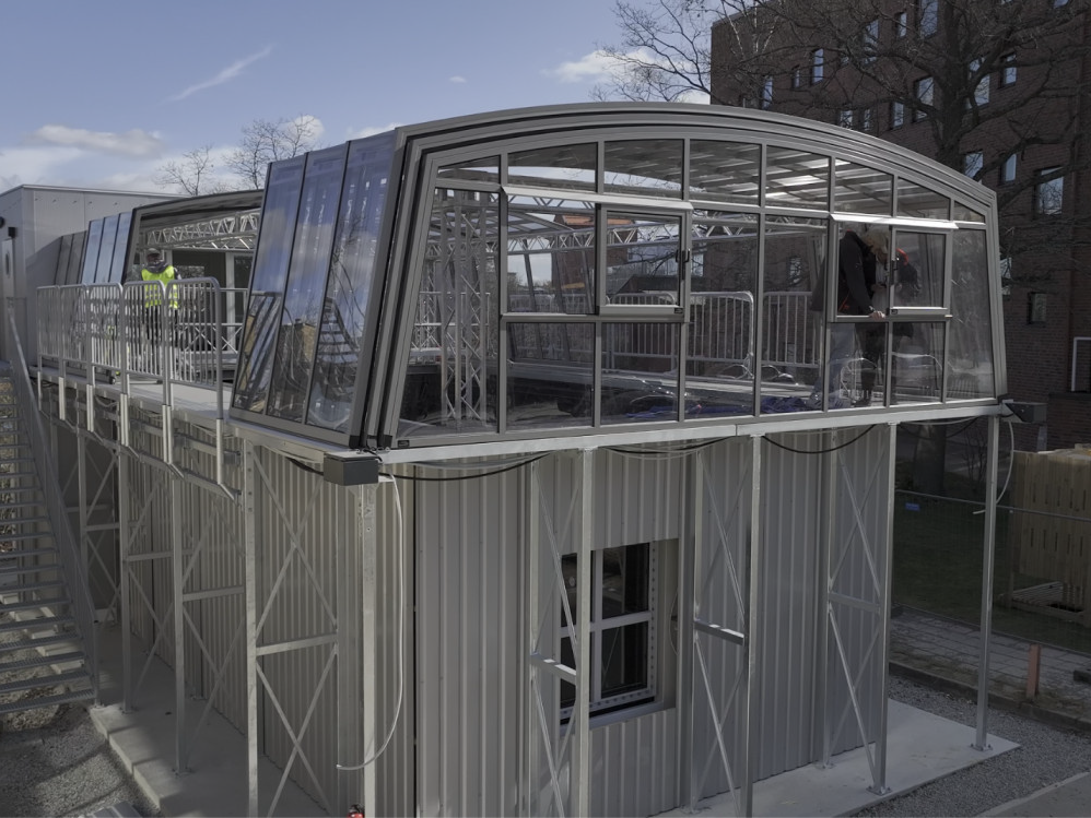 A modern, two-storey building with a curved glass and metal upper level and metal lower level. Two people stand on the upper deck, and an outdoor staircase leads to the roof. A brick building is visible in the background.