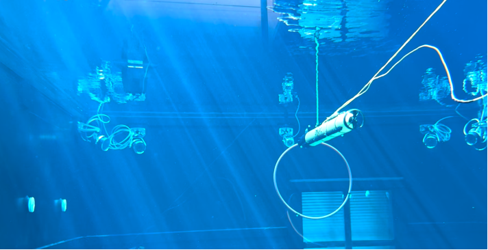 A cylindrical underwater robot is suspended by a cable in a blue-lit tank, surrounded by cameras and sensors attached to the tank walls, with light rays streaming through the water.