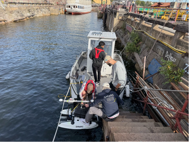 Four people prepare equipment on a small boat moored by a stone quay. Two wear SMARC jackets, and one wears a red life jacket. The water is calm and industrial buildings are visible in the background.
