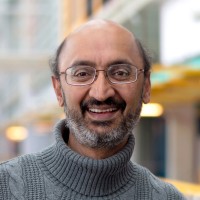 A smiling man with glasses, a balding head, and a beard, wearing a grey polo neck jumper, stands in a softly blurred indoor setting with warm lighting.