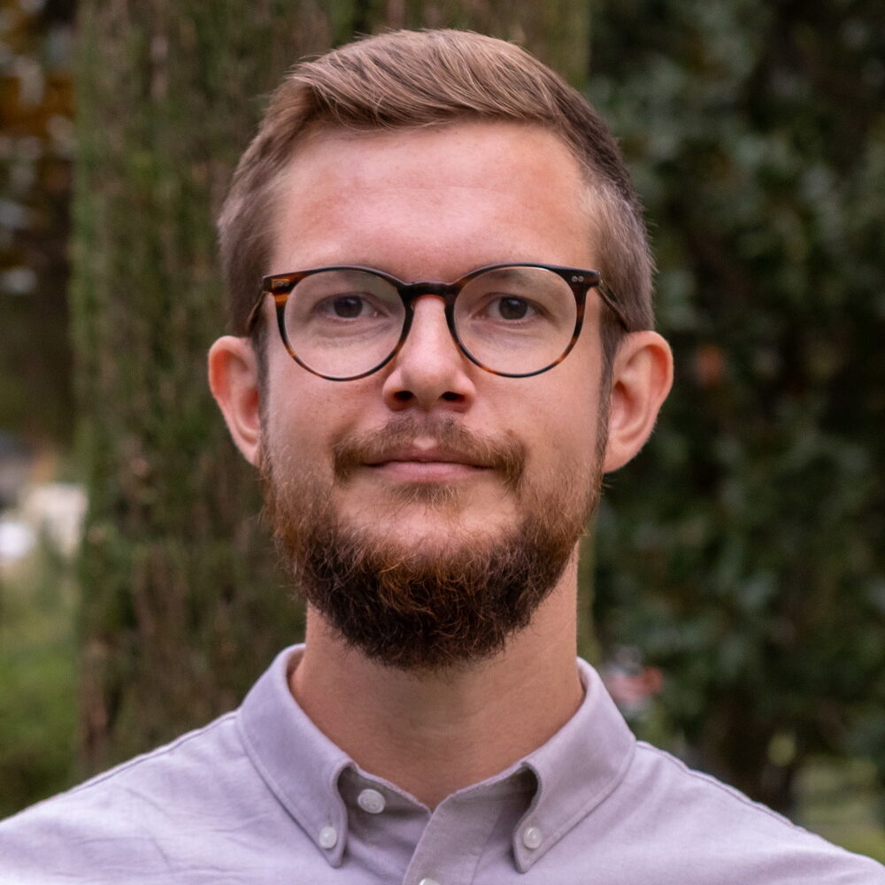 A young man with short light brown hair, a beard, and glasses, wearing a light grey button-up shirt, stands outdoors in front of greenery and trees, looking at the camera with a neutral expression.